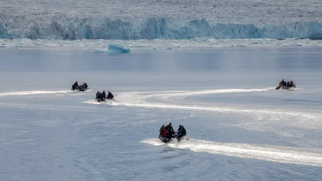 Four small inflatable boats full of passengers leave white wakes across a calm fjord towards a glacier front scattered with ice