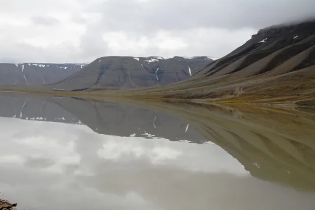 The still, grey water of Isdammen reflecting the brown mountainsides of Adventdalen under a cloudy sky
